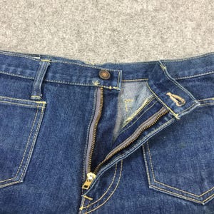 May include: Close-up of blue denim jeans with a partially unzipped brass zipper. The jeans have yellow stitching, a button closure, and front pockets. The fabric is a medium wash denim.