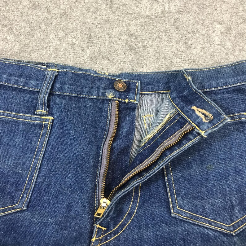 May include: Close-up of blue denim jeans with a partially unzipped brass zipper. The jeans have yellow stitching, a button closure, and front pockets. The fabric is a medium wash denim.