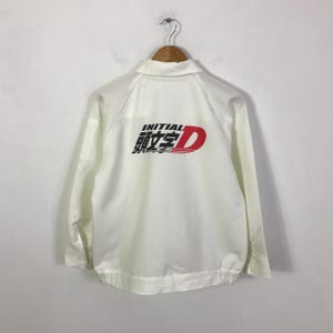 Puede incluir: Chaqueta blanca con cuello y mangas largas. La parte trasera de la chaqueta presenta el texto "INITIAL" en negro y caracteres japoneses, con una gran "D" roja a la derecha. La chaqueta está colgada de una percha de madera.