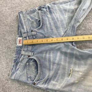 May include: Light blue denim jeans with a distressed look. The jeans have a classic five-pocket design, a button and zipper closure, and a small brand label. A wooden ruler is placed across the jeans, measuring approximately 13 inches.