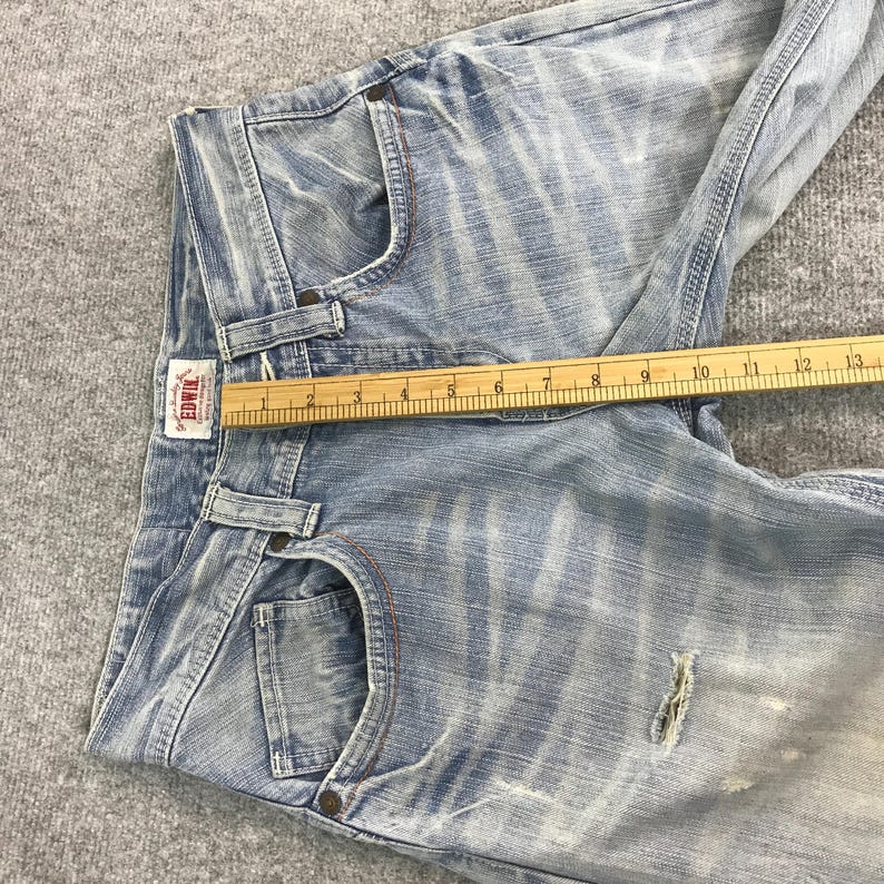 May include: Light blue denim jeans with a distressed look. The jeans have a classic five-pocket design, a button and zipper closure, and a small brand label. A wooden ruler is placed across the jeans, measuring approximately 13 inches.