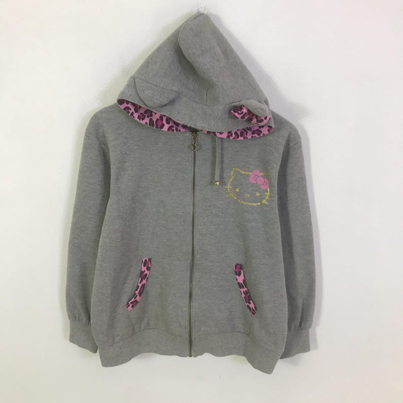 May include: A gray zip-up hoodie featuring a Hello Kitty design with a pink bow. The hood has cat ears and a pink leopard print lining. The pockets also have pink leopard print accents.
