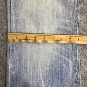 May include: Close-up of light blue denim fabric with a wooden ruler measuring across the material. The ruler shows measurements in inches, with markings from 1 to 9 inches.