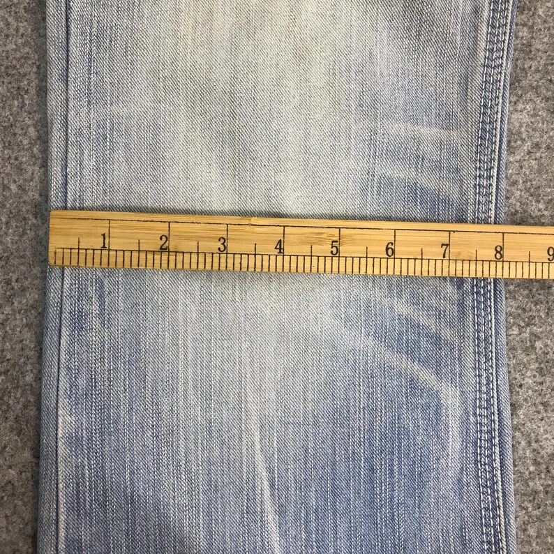 May include: Close-up of light blue denim fabric with a wooden ruler measuring across the material. The ruler shows measurements in inches, with markings from 1 to 9 inches.