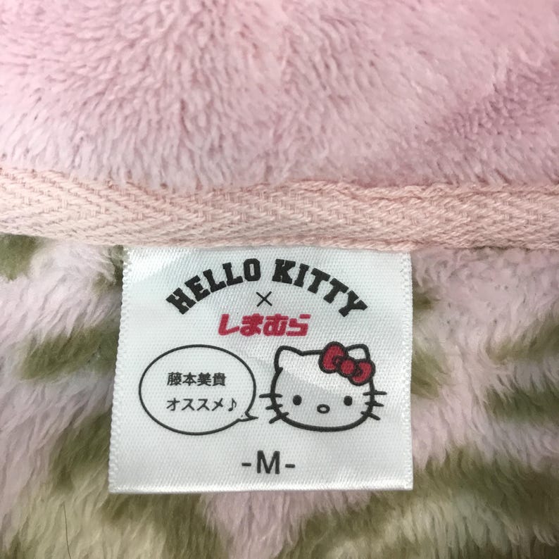 Puede incluir: Primer plano de una etiqueta blanca con el logotipo de Hello Kitty y texto en japon&eacute;s. La etiqueta presenta una imagen de dibujos animados de Hello Kitty. El fondo es rosa y verde, con un ribete rosa.