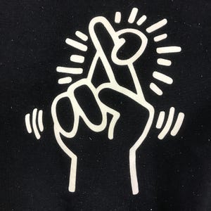 K&ouml;nnte beinhalten: Schwarze Grafik mit einer wei&szlig;en Hand, die ein gekreuztes Fingerzeichen macht, umgeben von strahlenf&ouml;rmigen und gebogenen Linien. Das Design ist eine einfache, k&uuml;hne Illustration auf schwarzem Hintergrund.