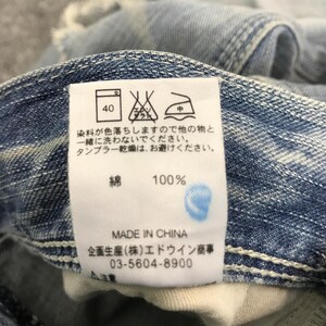 May include: Close-up of a white tag with Japanese text and care symbols, attached to a pair of light blue denim jeans. The tag indicates the fabric is 100% cotton and made in China.