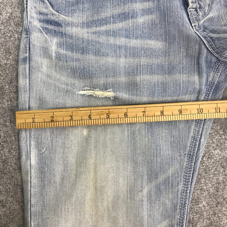 May include: Light blue denim jeans with a distressed design, featuring a wooden ruler measuring approximately 10 inches. The jeans have a faded appearance and a small tear near the knee, showcasing a worn aesthetic.