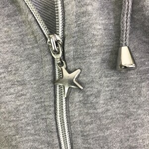 May include: Close-up of a gray hooded sweatshirt with a silver zipper and a star-shaped charm. The hood's drawstring has silver tips. The fabric is a heathered gray.