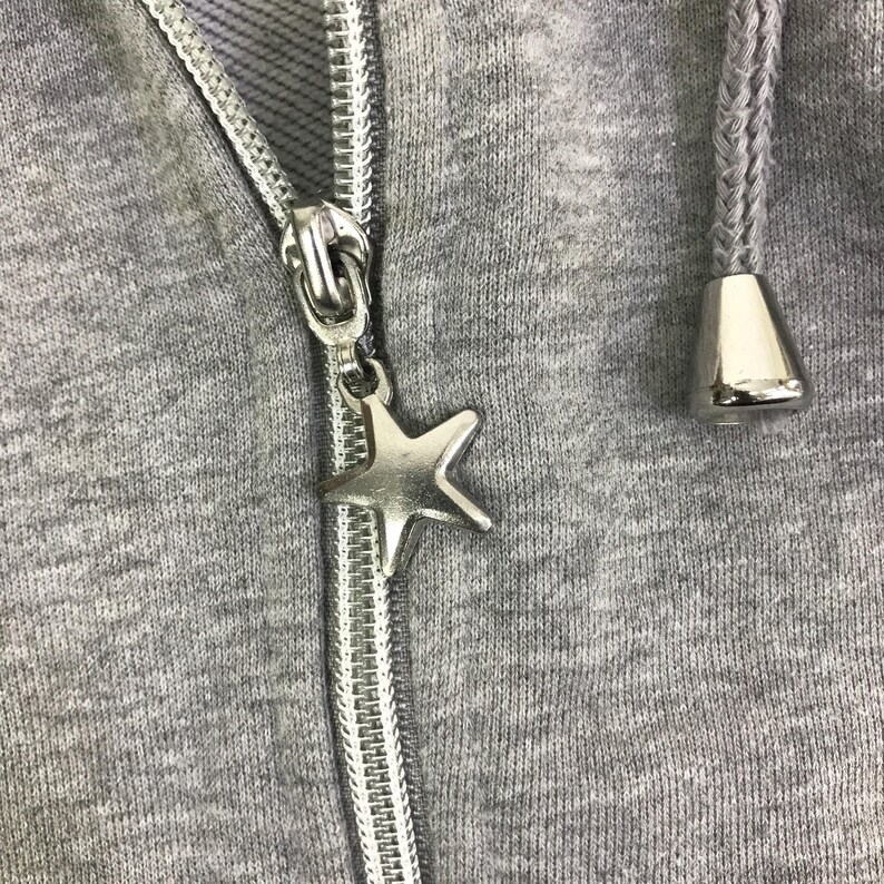 May include: Close-up of a gray hooded sweatshirt with a silver zipper and a star-shaped charm. The hood's drawstring has silver tips. The fabric is a heathered gray.