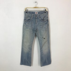 May include: Light blue denim jeans with a distressed look, featuring a faded wash and a small rip on the right leg. The jeans are hanging on a gold-colored hanger. The brand "Edwin" is visible on the front.