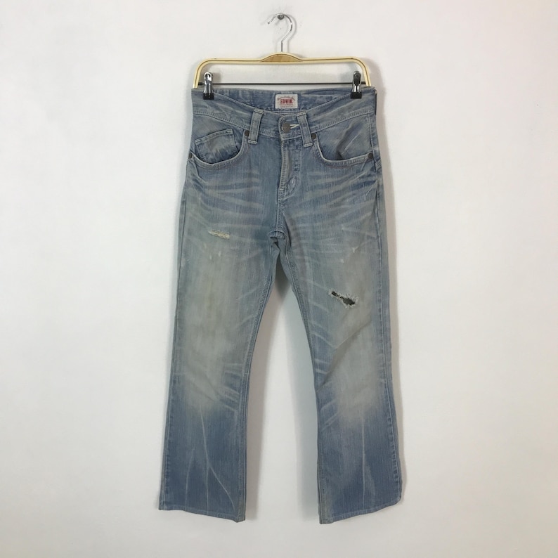 May include: Light blue denim jeans with a distressed look, featuring a faded wash and a small rip on the right leg. The jeans are hanging on a gold-colored hanger. The brand "Edwin" is visible on the front.