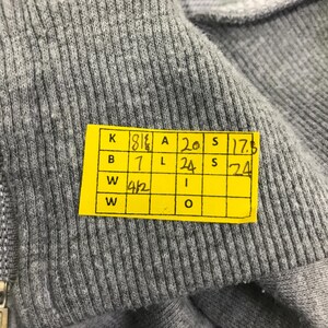 May include: Close-up of a gray, ribbed fabric. A yellow tag with black text is attached to the fabric. The tag contains numbers and letters in a grid format. The fabric appears to be part of a garment.