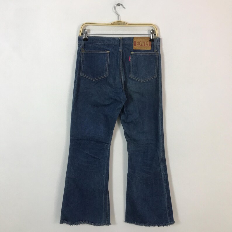 May include: Dark blue denim jeans with a flared leg design, featuring two back pockets and a leather patch with the brand name "EDWIN". The jeans are hanging on a metal hanger.