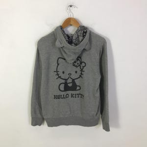 May include: A gray hooded sweatshirt featuring a black Hello Kitty graphic on the back. The hood has cat ears and a patterned lining. The words "HELLO KITTY" are printed below the graphic.
