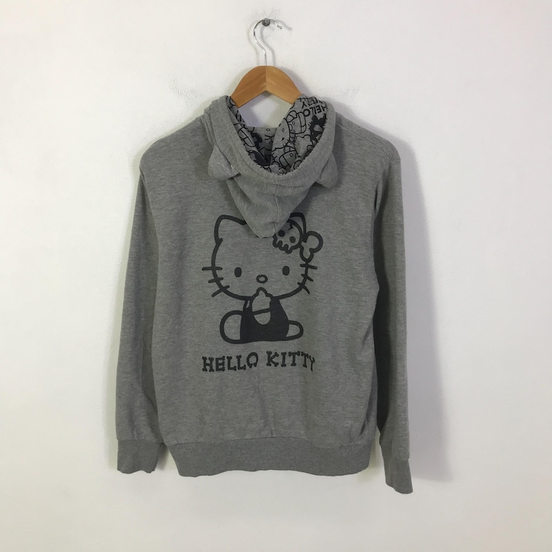 May include: A gray hooded sweatshirt featuring a black Hello Kitty graphic on the back. The hood has cat ears and a patterned lining. The words "HELLO KITTY" are printed below the graphic.