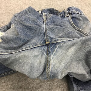 May include: Close-up of light blue denim jeans, showing the back pockets, belt loops, and stitching. The jeans have a distressed look with some fading and wear. The fabric is folded, revealing the texture and construction details.