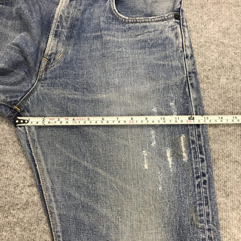 May include: Close-up of blue denim jeans with a measuring tape showing a length of approximately 12 inches. The jeans have distressed details and a faded appearance, with visible wear and tear.