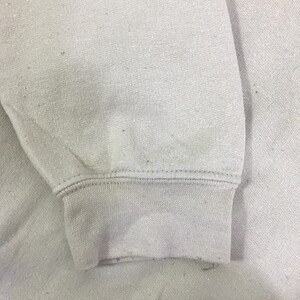May include: Close-up of a pale grey sweatshirt sleeve. The cuff is ribbed and the fabric has a slightly textured appearance. The stitching is visible along the cuff and sleeve seam.