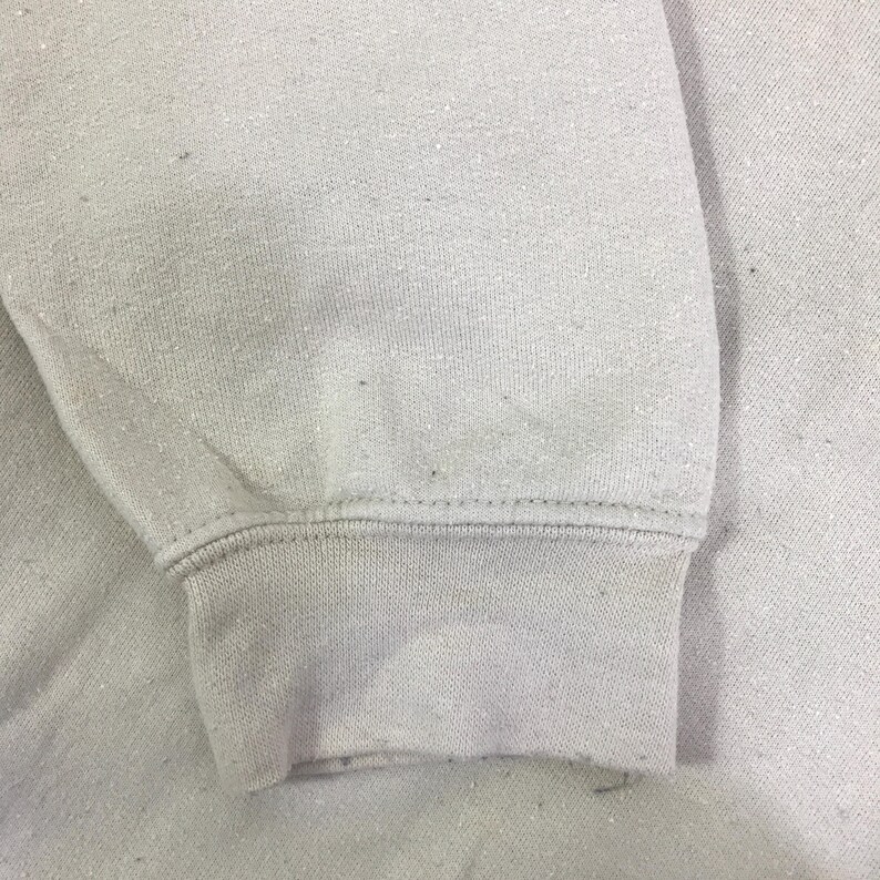 May include: Close-up of a pale grey sweatshirt sleeve. The cuff is ribbed and the fabric has a slightly textured appearance. The stitching is visible along the cuff and sleeve seam.