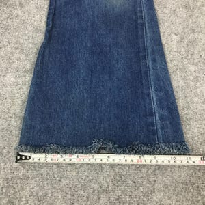 May include: Close-up of the bottom of a pair of blue denim jeans with a frayed hem. A measuring tape is placed across the bottom of the jeans, showing measurements in inches. The jeans have a classic, casual style.