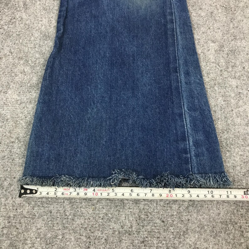May include: Close-up of the bottom of a pair of blue denim jeans with a frayed hem. A measuring tape is placed across the bottom of the jeans, showing measurements in inches. The jeans have a classic, casual style.
