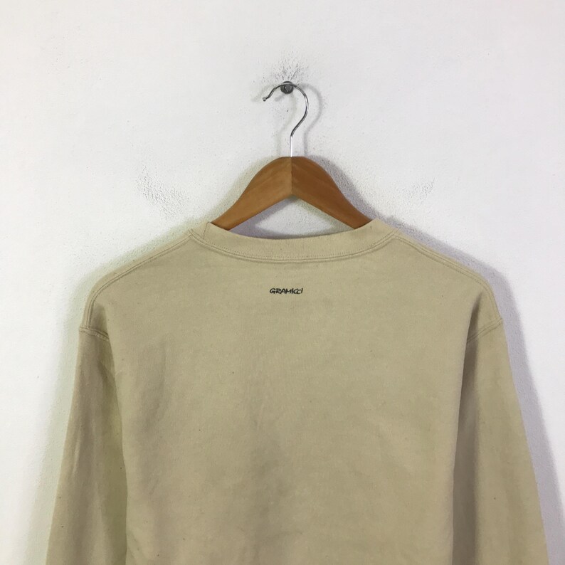 May include: A beige sweatshirt hangs from a wooden hanger against a white wall. The sweatshirt has a crew neck and long sleeves. The brand name "GRAMICCI" is printed in small, dark letters on the back of the collar.