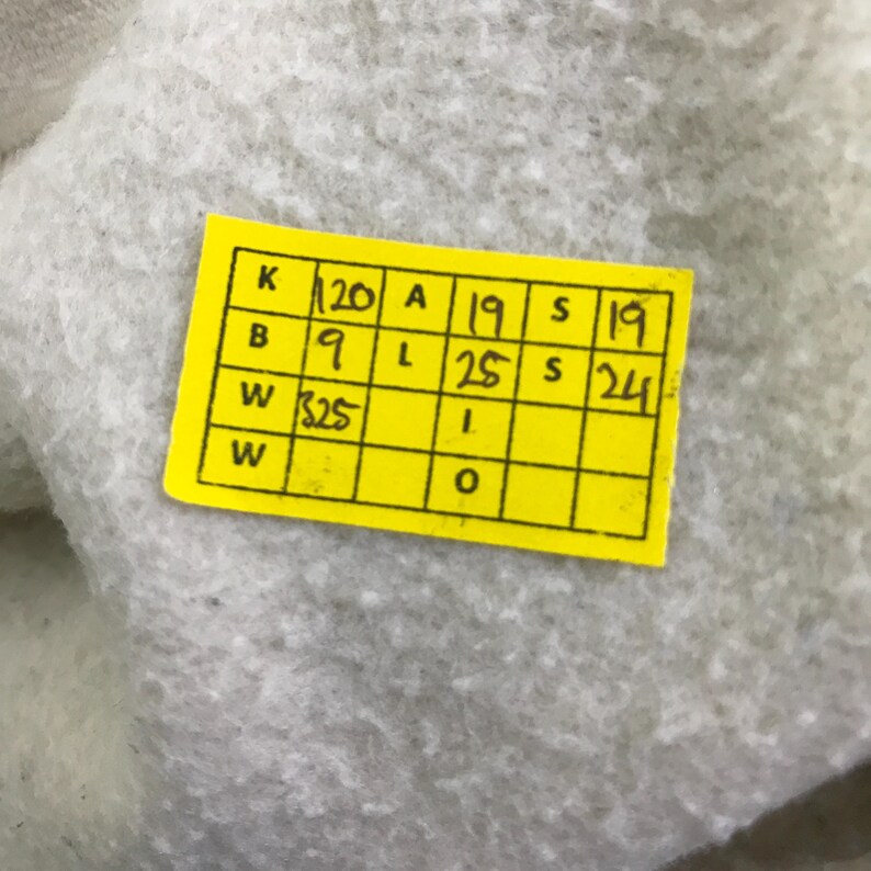 May include: A yellow rectangular tag with black text and a grid of boxes, resting on a light grey, textured surface. The text includes numbers and letters. The image is a close-up.