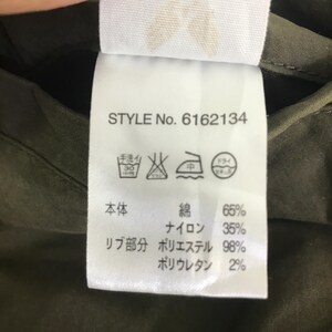 May include: A white garment care label with black text reading STYLE No. 6162134, featuring laundry symbols and material composition percentages for the main body and ribbing, attached to an olive green fabric background.