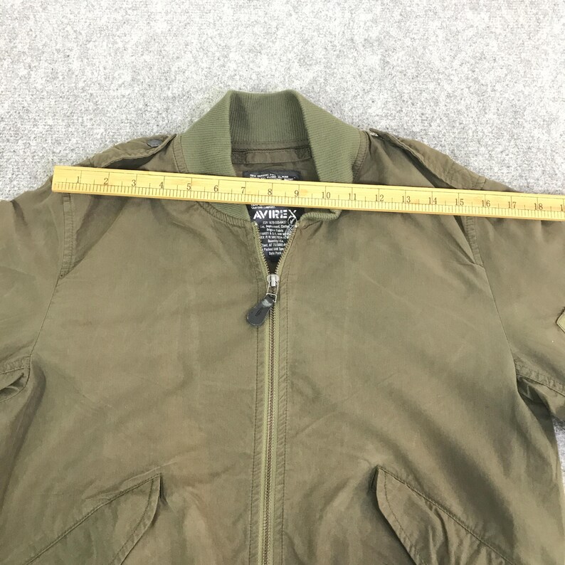 May include: An olive green bomber jacket with a ribbed collar, a central zipper closure, and two front pockets. A yellow wooden ruler is placed horizontally across the chest, showing a measurement of approximately 18 inches across the jacket.