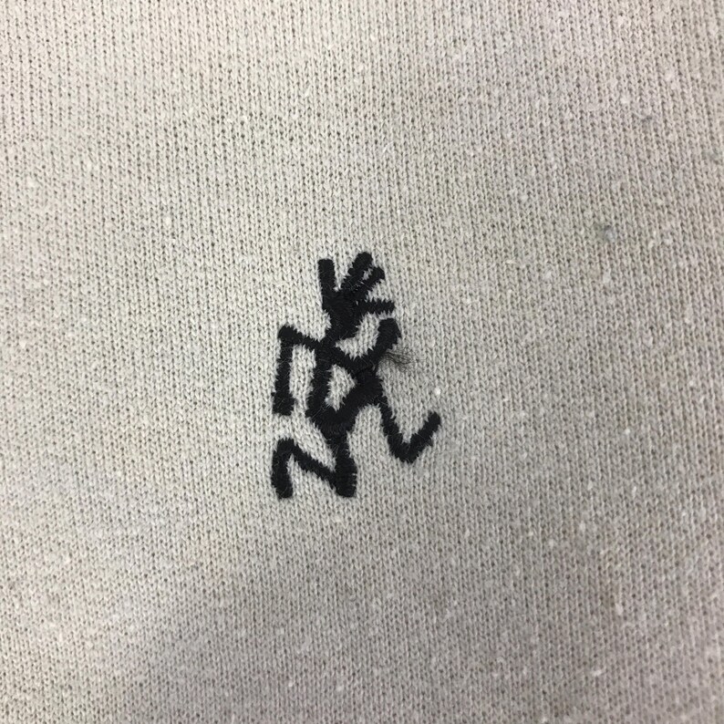 May include: Close-up of a light grey fabric featuring a black embroidered figure. The figure is a stylized representation of a person in motion. The fabric has a textured appearance with small white flecks.