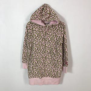 Puede incluir: Vestido sudadera con capucha rosa con estampado de leopardo en tonos marr&oacute;n y amarillo. El vestido tiene capucha con cord&oacute;n, mangas largas y dobladillo y pu&ntilde;os de canal&eacute; rosas. Un peque&ntilde;o logo es visible en la manga.