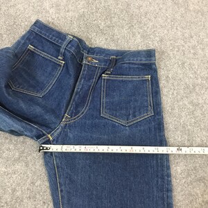 May include: Close-up of blue denim jeans with yellow stitching and a button closure. A measuring tape shows the jeans are approximately 13 inches wide. The jeans have two front pockets and two back pockets.