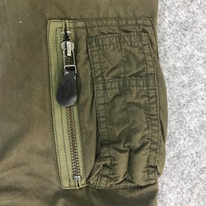 May include: An olive green garment featuring a vertical zippered pocket with a dark brown leather-like pull tab. The pocket is positioned next to a rectangular patch pocket with visible vertical stitching, constructed from a durable fabric.