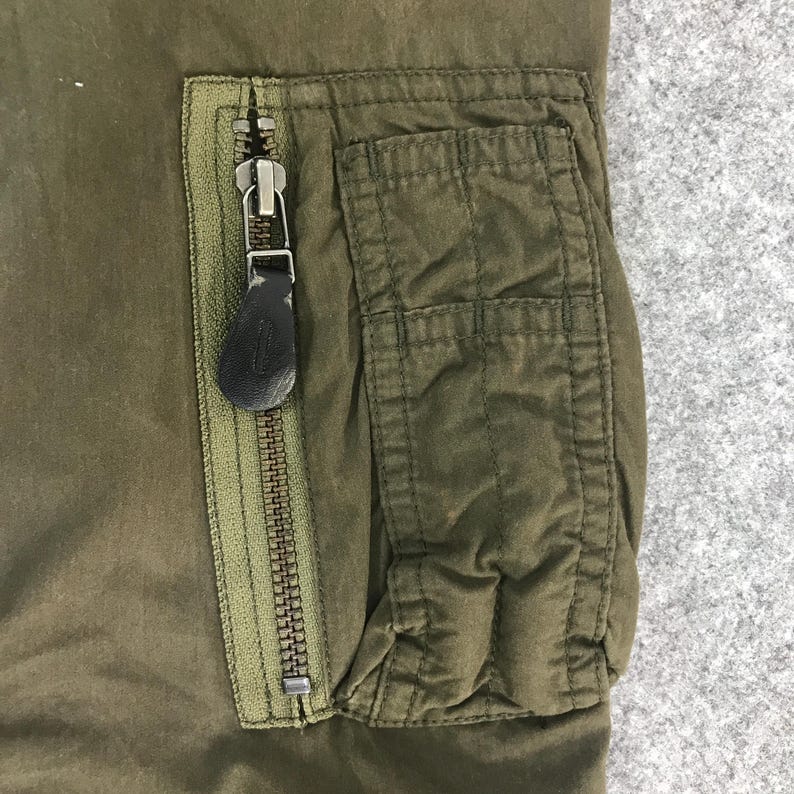 May include: An olive green garment featuring a vertical zippered pocket with a dark brown leather-like pull tab. The pocket is positioned next to a rectangular patch pocket with visible vertical stitching, constructed from a durable fabric.