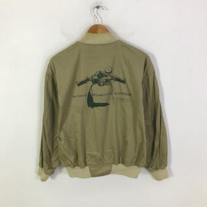 May include: Khaki bomber jacket with a cream-coloured collar, cuffs, and hem. The back displays a green motorcycle graphic and the words "ALWAYS DELIGHTFUL SURPRISE". The jacket is suspended from a wooden hanger.