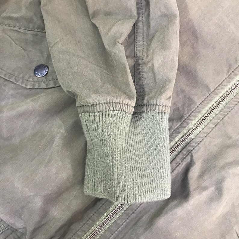 May include: An olive green jacket featuring a ribbed knit cuff, a long sleeve with visible stitching, and a metal zipper closure. A pocket flap with a dark circular button is positioned on the upper left side of the garment, showing fabric texture.