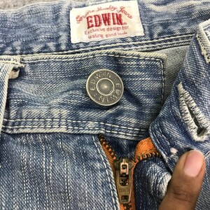 May include: Close-up of light blue denim jeans with a metal button and zipper. The jeans have white stitching and a label that reads "Genuine Quality Jeans EDWIN Exclusive design for lasting good look."