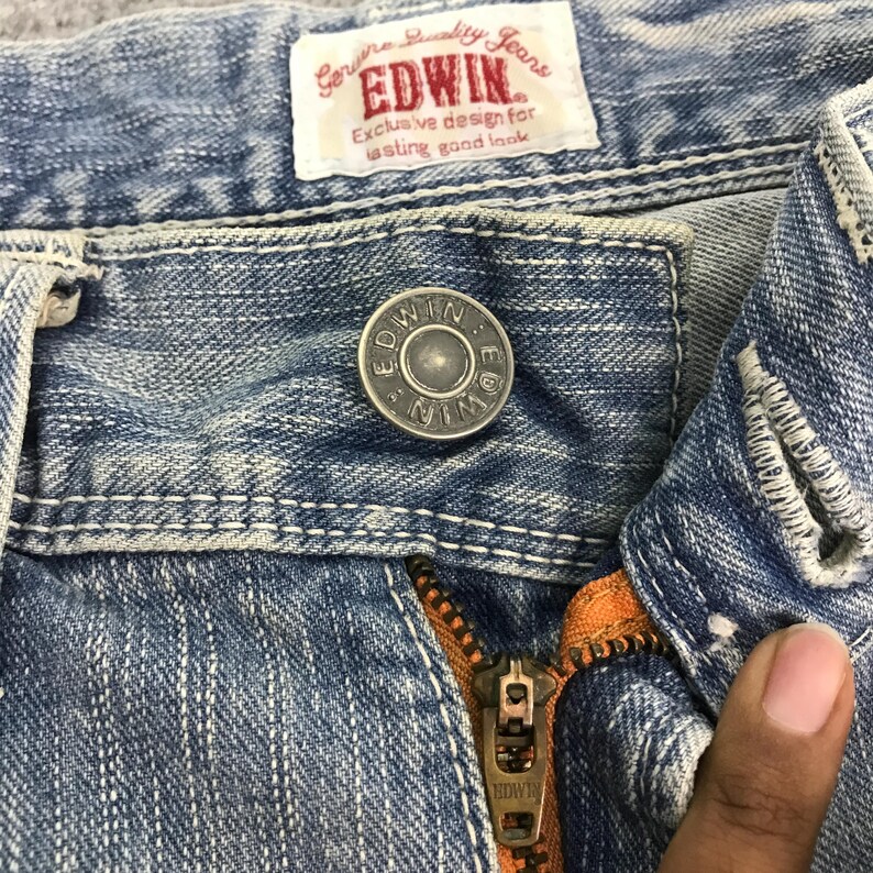 May include: Close-up of light blue denim jeans with a metal button and zipper. The jeans have white stitching and a label that reads "Genuine Quality Jeans EDWIN Exclusive design for lasting good look."