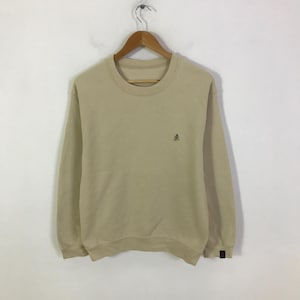 May include: A beige sweatshirt with a crew neck and long sleeves. A small, dark emblem is on the left chest. The sweatshirt is suspended from a wooden hanger against a white backdrop. The cuffs have a small, dark brown patch.