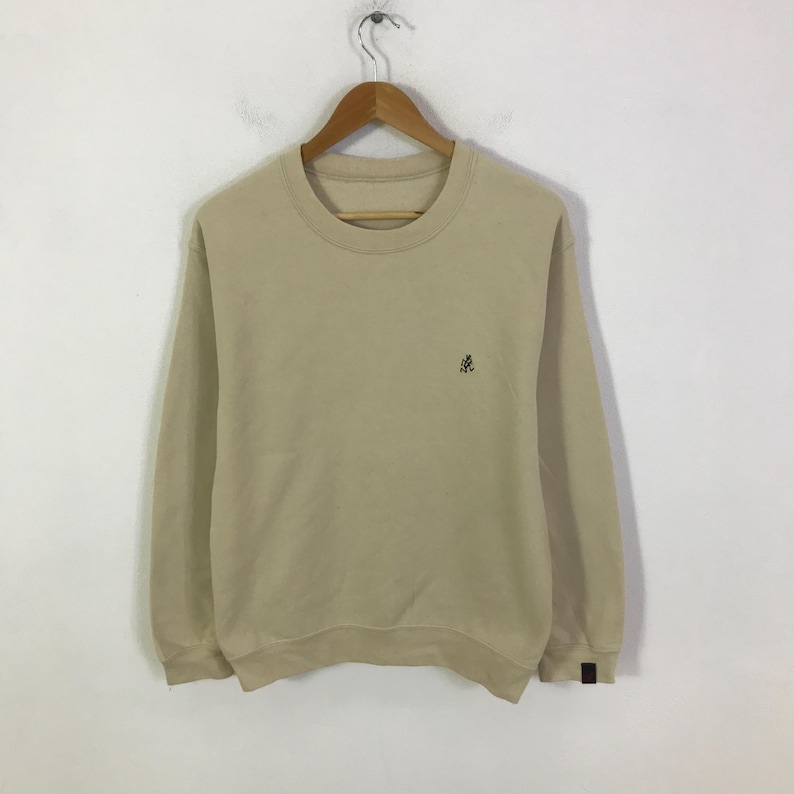 May include: A beige sweatshirt with a crew neck and long sleeves. A small, dark emblem is on the left chest. The sweatshirt is suspended from a wooden hanger against a white backdrop. The cuffs have a small, dark brown patch.