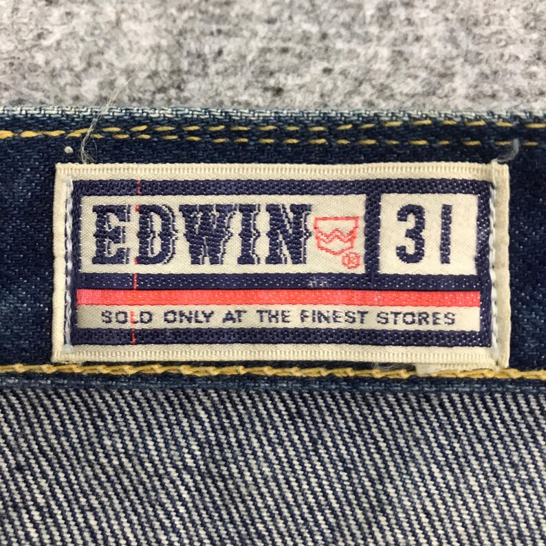 May include: Close-up of a denim jeans label. The label is rectangular with the brand name "EDWIN" in white letters. It also features the number "31" and the text "SOLD ONLY AT THE FINEST STORES". The label has a red and white design.