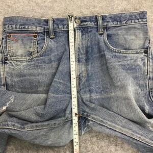 May include: Light blue denim jeans with a faded wash. The jeans have a classic five-pocket design, a button and zipper closure, and a straight leg. The jeans are being measured with a tape measure, showing a length of approximately 15 inches.