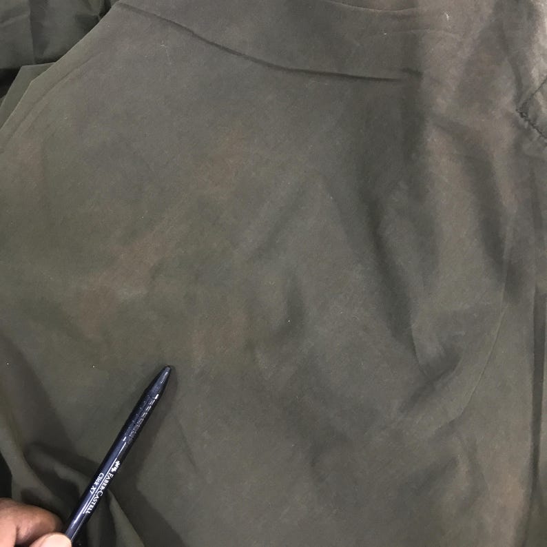 May include: A dark olive green fabric with a smooth, matte texture. A dark blue Faber-Castell Click X7 ballpoint pen is positioned diagonally across the lower left portion of the textile, showing the brand name and model printed on the barrel.