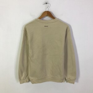 May include: A tan sweatshirt with long sleeves hangs from a wooden hanger. The word "GRAMICCI" is printed in black on the back of the sweatshirt. The sweatshirt is made of a soft, comfortable material.