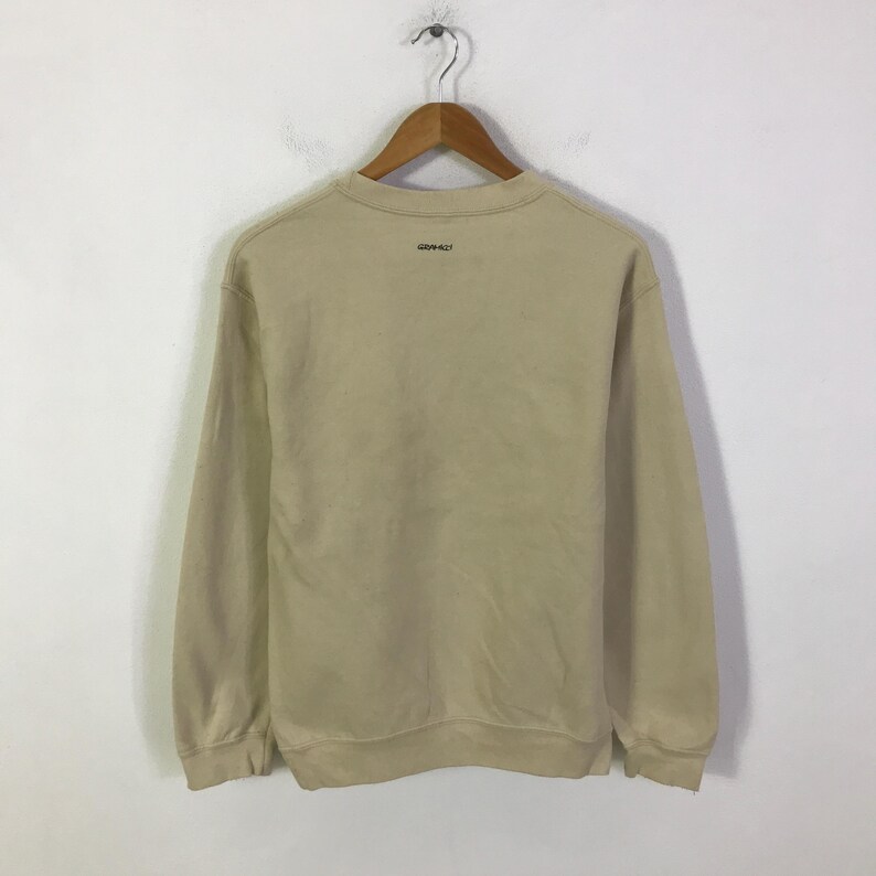 May include: A tan sweatshirt with long sleeves hangs from a wooden hanger. The word "GRAMICCI" is printed in black on the back of the sweatshirt. The sweatshirt is made of a soft, comfortable material.