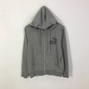 May include: A gray zip-up hoodie featuring the Hello Kitty logo in black on the front. The hoodie has a drawstring hood, a front pocket, and ribbed cuffs and hem. The zipper is silver.