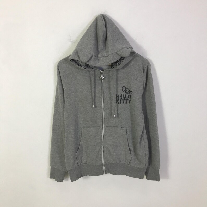 May include: A gray zip-up hoodie featuring the Hello Kitty logo in black on the front. The hoodie has a drawstring hood, a front pocket, and ribbed cuffs and hem. The zipper is silver.