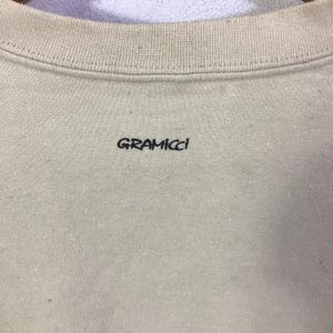 May include: A close-up of a beige sweatshirt with a ribbed crew neck. The black text "GRAMICCI" is printed on the front. The fabric has a slightly textured appearance.