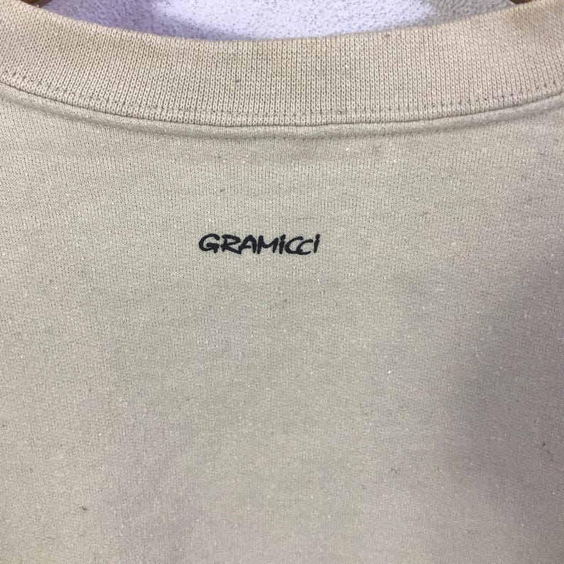 May include: A close-up of a beige sweatshirt with a ribbed crew neck. The black text "GRAMICCI" is printed on the front. The fabric has a slightly textured appearance.